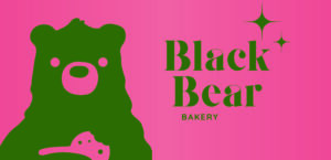 A bright pink background (HEx#E4747C) with a green bear eating a cookie. A logo off to the right side says "Black Bear Bakery" and has two stars over the right top corner. New Kitchen and Market Update for March 2024